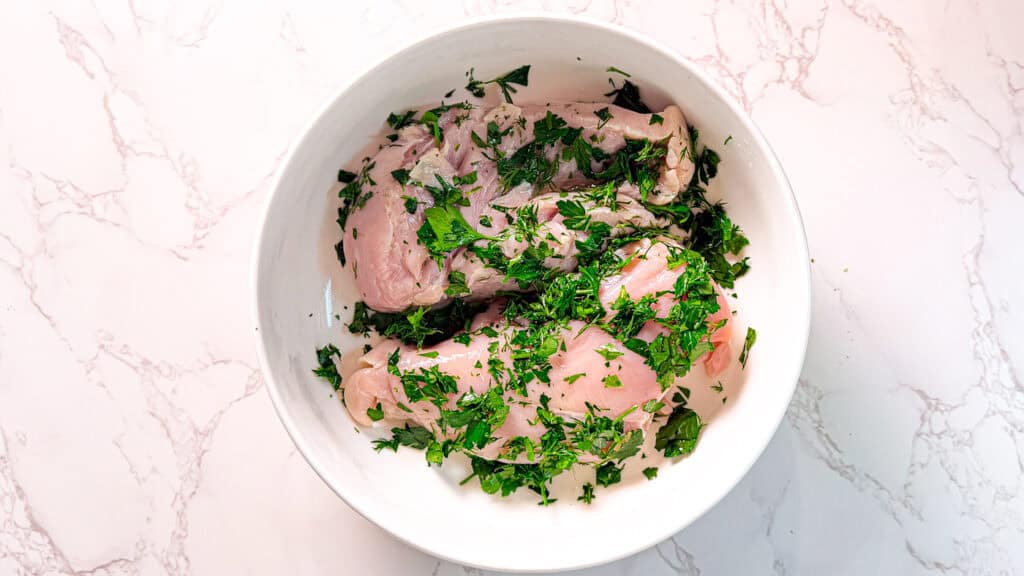 Juicy Lemon herb chicken process shot marinade, raw chicken breast with parsley and lemon, in a white bowl on a white marble surface