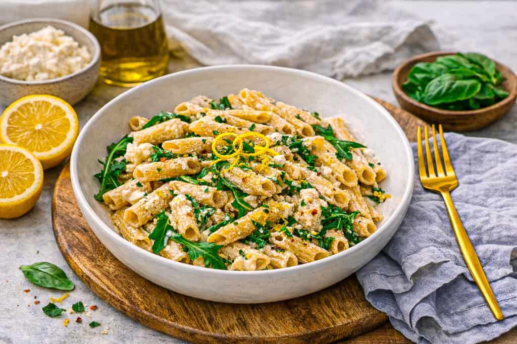 Lemon ricotta protein pasta, penne pasta with ricotta sauce and arugula on a concrete surface with a blue linen napkin and a gold fork, in the background is a lemon, a jar of olive oil, a small bowl of basil, and a small bowl of ricotta cheese.