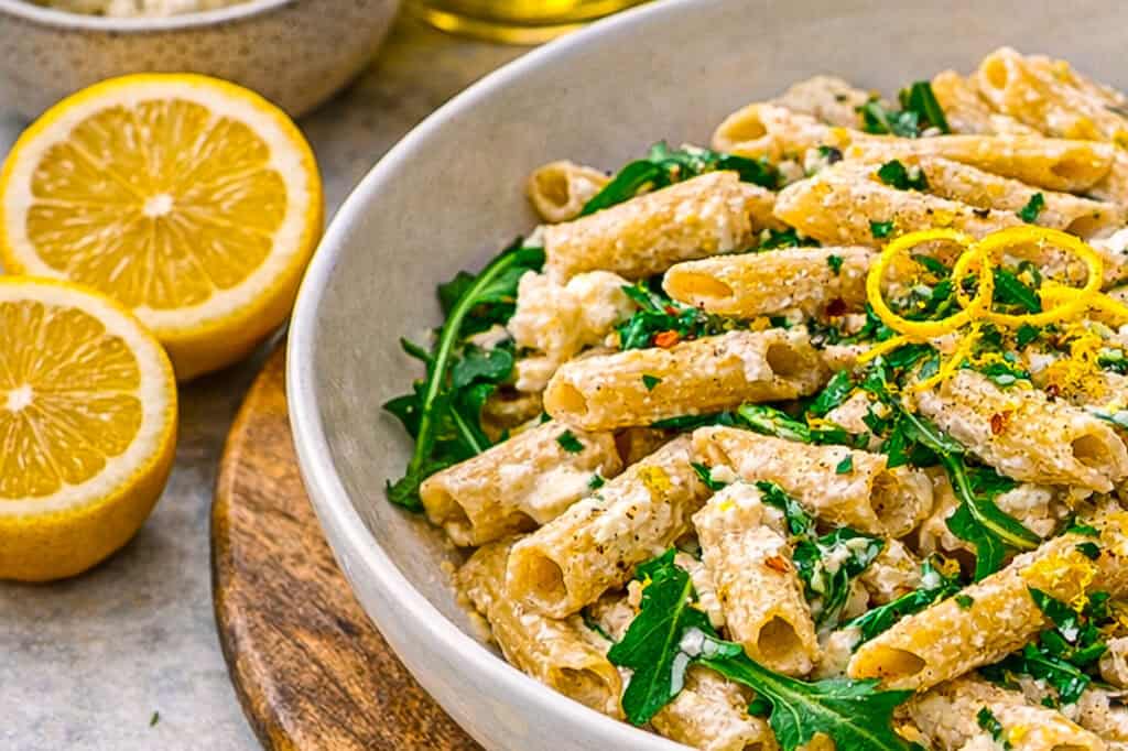 Lemon ricotta protein pasta, penne pasta with ricotta sauce and arugula on a concrete surface with a blue linen napkin and a gold fork, in the background is a lemon, and a small bowl of ricotta cheese.