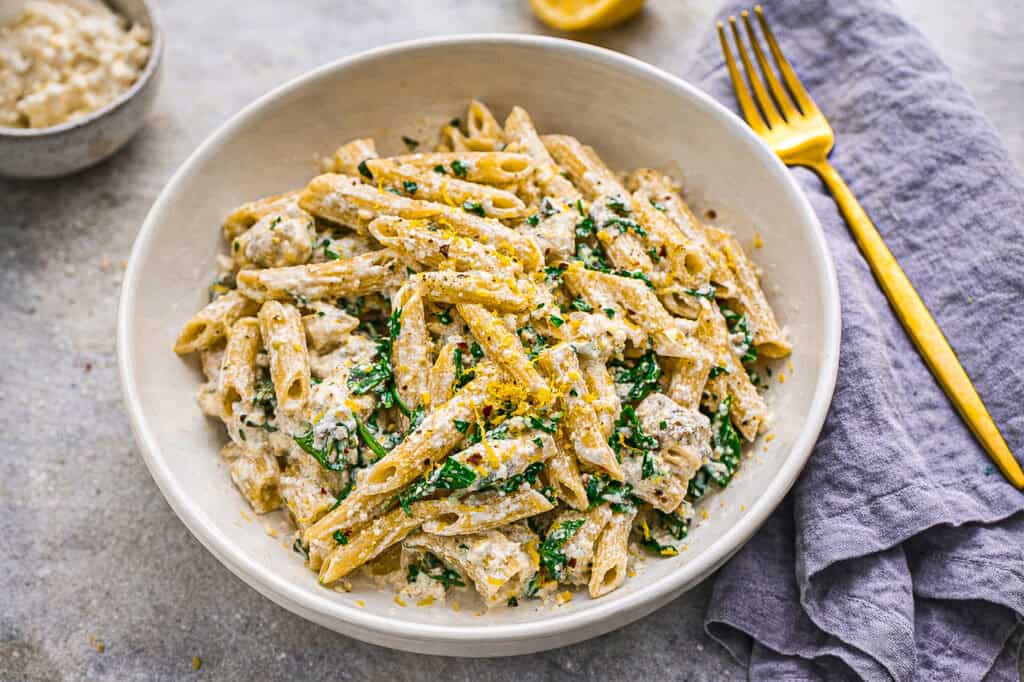 Lemon ricotta protein pasta, penne pasta with ricotta sauce and arugula on a concrete surface with a blue linen napkin and a gold fork, in the background is a lemon, and a small bowl of ricotta cheese.
