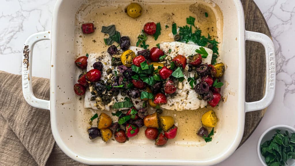 Lemon garlic baked cod with tomatoes, parsley, basil, olives, capers, Greek oregano, lemon, and spices in a baking dish after being cooked.
