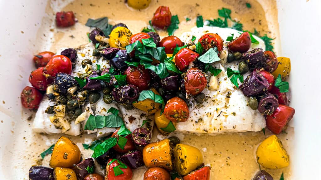 Herb crusted baked cod in a baking dish with olive oil, lemon, cherry tomatoes, fresh herbs