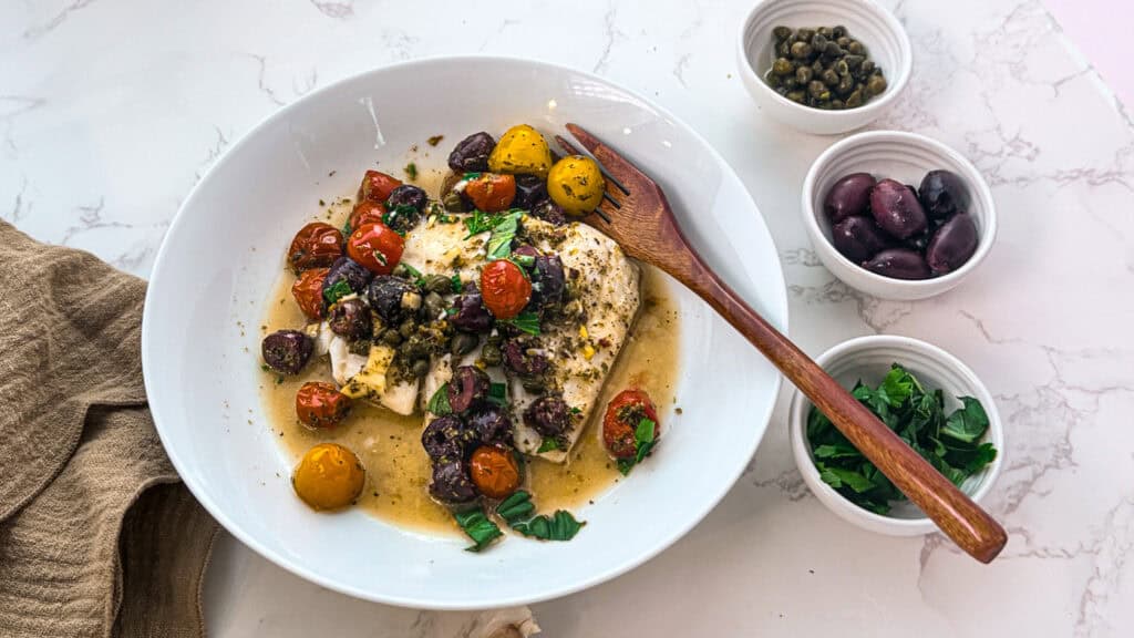 cod with capers and lemon in a white  bowl with three small bowls with capers, olives, and fresh herbs with a wooden fork and an oatmeal linen napkin on. white marble.