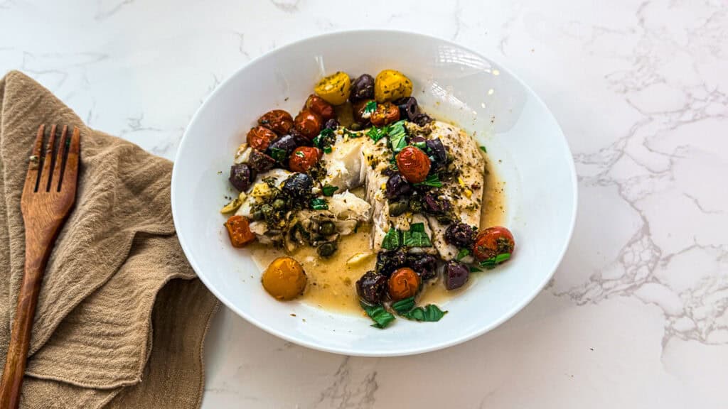 Cod with cherry tomatoes and herbs in a white bowl with an oatmeal linen napkin and a wooden fork on top of white marble.