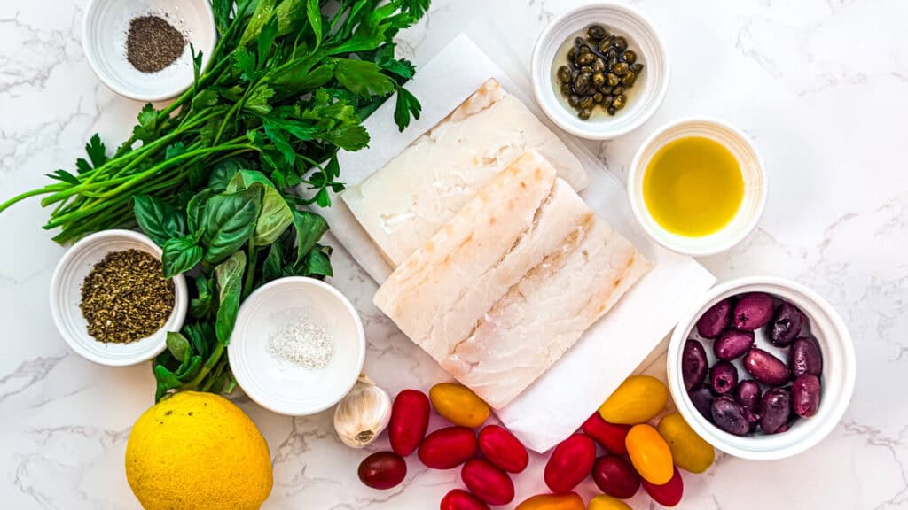 Baked cod with tomatoes and olives, an ingredients flatlay with parsley, basil, black pepper, Greek oregano, code, capers, olive oil, olives, cherry tomatoes, and garlic.