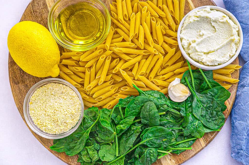 Lemon Ricotta protein pasta, ingredients flatlay with protein penne, lemon, spinach, garlic, ricotta, olive oil, and parmesan cheese on a cutting board with a blue linen napkins on a white surface