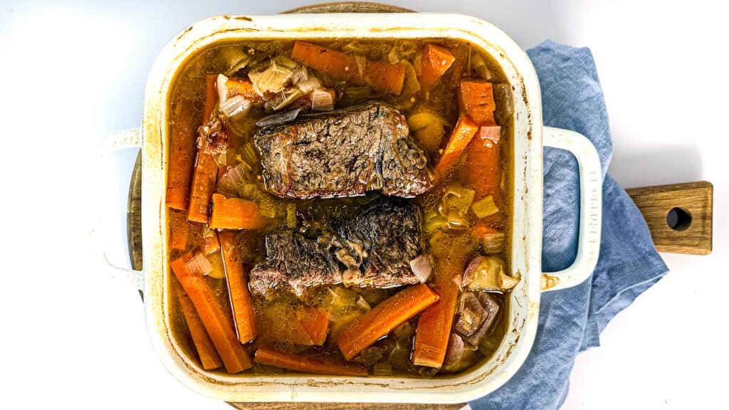 tender pot roast with carrots and onions, pot roast, carrots, onions, leeks, parsnips, in chicken broth in a baking dish on a cutting board with a blue linen napkin on a white surface.