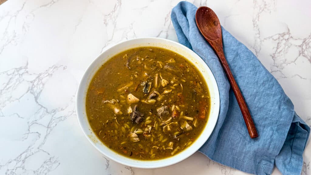 Chicken orzo soup in a  white bowl, with a blue linen napkins, a wooden spoon, sitting on a white marble surface.