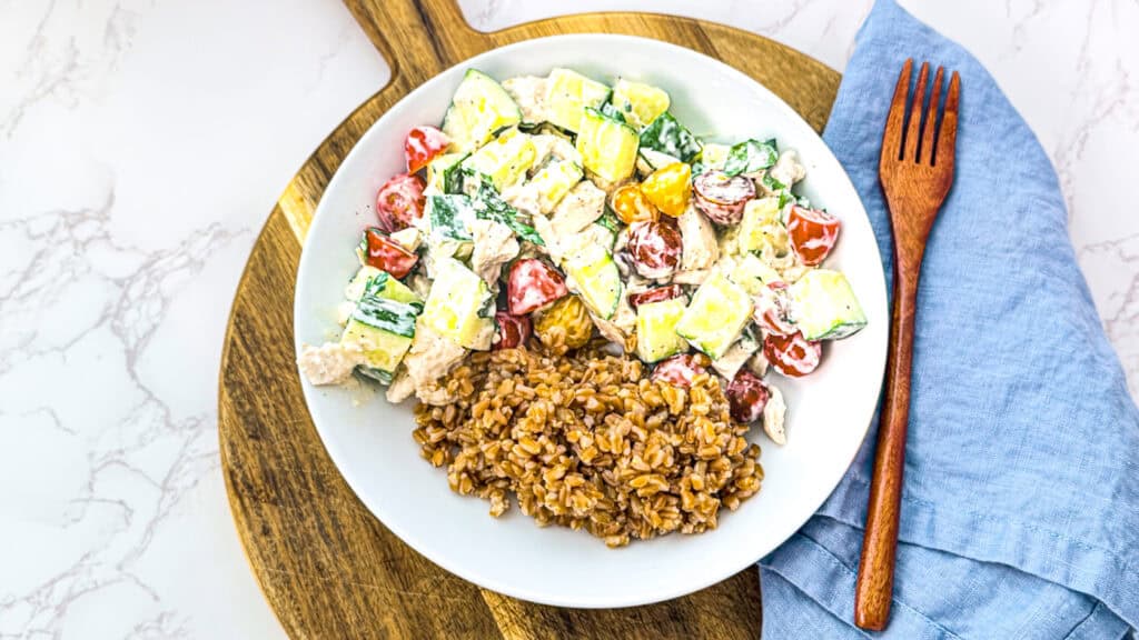 lemon herb chicken bowl with feat, cucumbers, parsley, cherry tomatoes, chicken, and yogurt dressing in a wet bowl on a cutting board with a  blue linen napkin and a wooden fork.