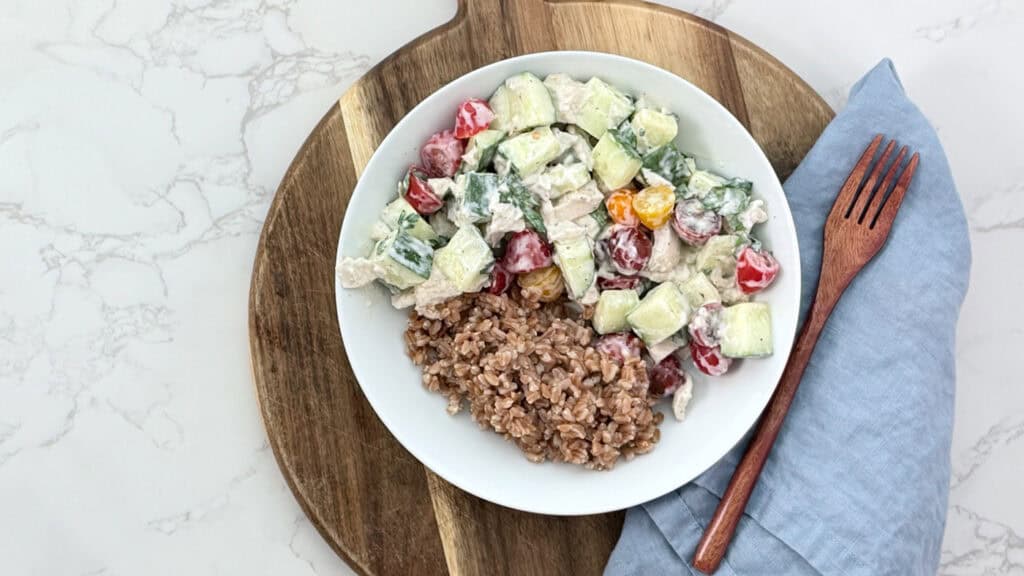 chicken salad with cucumber, cherry tomatoes and farro with a yogurt dressing on a cutting board, with a blue linen napkin and a wooden fork