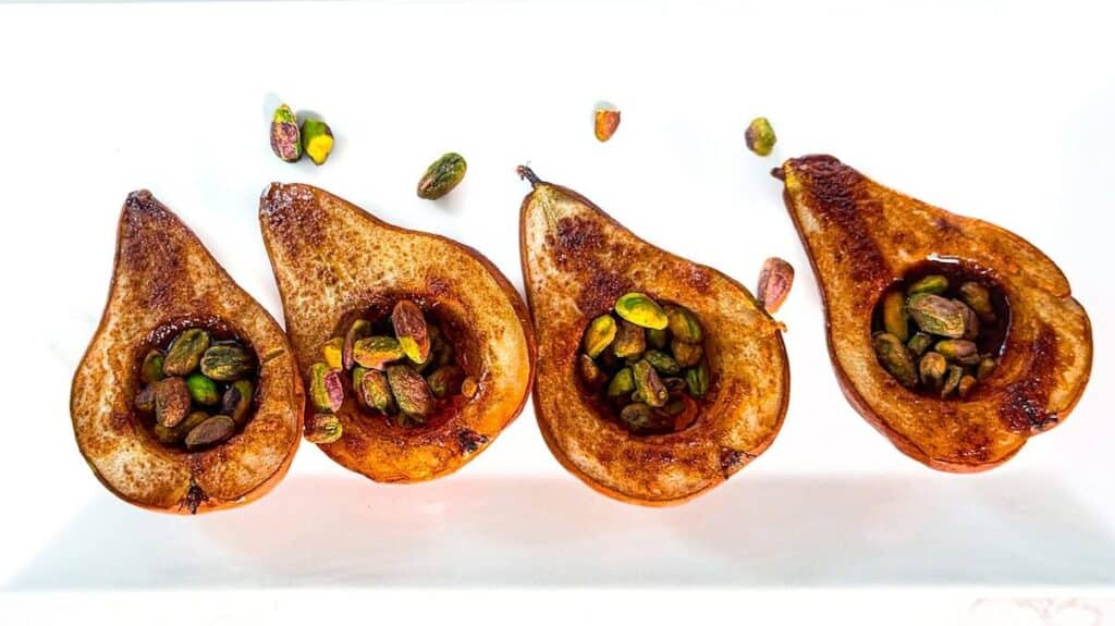 Four baked pears topped with chopped pistachios on a white plate.