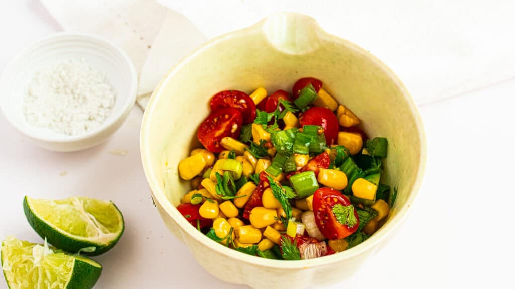Fresh corn, cherry tomatoes, and green onions in a white bowl, garnished with lime wedges.