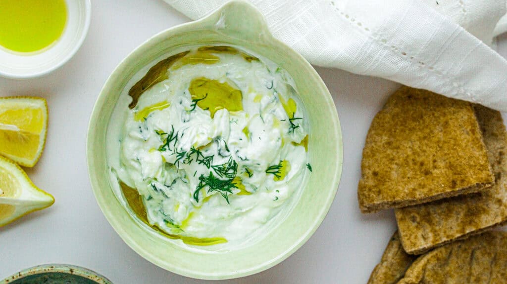 Creamy tzatziki sauce garnished with dill, served with pita bread and lemon wedges.