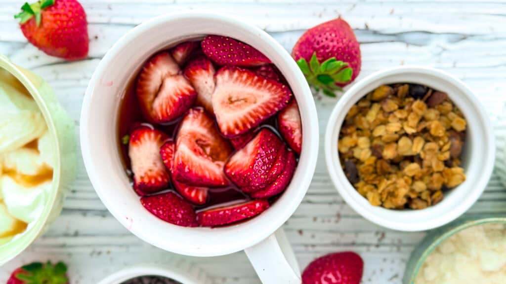 Bowl of sliced strawberries with fresh strawberries around on a white wooden surface.