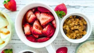 Bowl of sliced strawberries with fresh strawberries around on a white wooden surface.