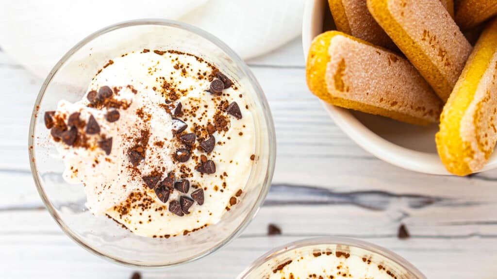Tiramisu yogurt cups topped with cocoa powder and chocolate chips, served with cookies on a white-wo.