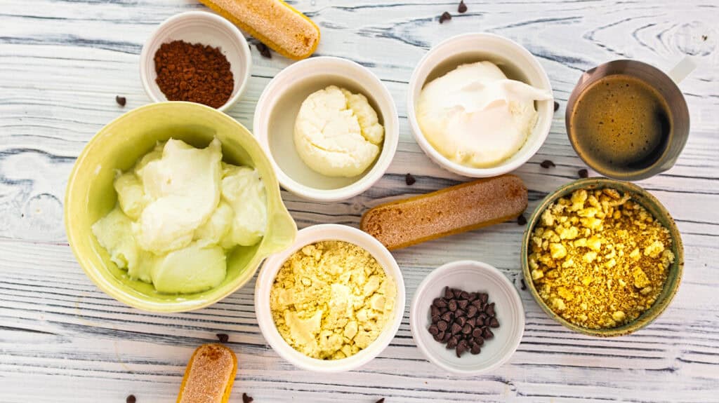Assorted ingredients for Tiramisu Yogurt Cups on a white wooden surface.