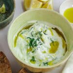Creamy tzatziki dip garnished with herbs, olive oil, and served with bread slices.