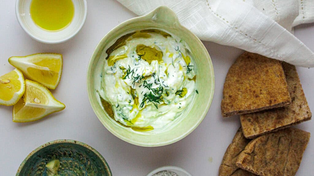 Creamy lemon herb High-Protein Greek Yogurt Tzatziki Dip garnished with dill, served with crispy crackers and lemon wedges.