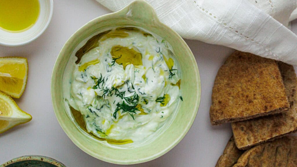 Delicious homemade High-Protein Greek Yogurt Tzatziki Dip garnished with dill, served with crispy pita chips and lemon wedges.